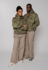 Deux mannequins portent des sweat-shirts à capuche assortis en motif camouflage vert, associés à des pantalons cargo beiges. Des baskets complètent les tenues.