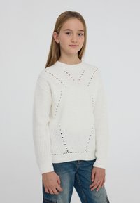 Biały sweter robiony na drutach z okrągłym dekoltem, z ozdobnymi wstawkami oraz ściągaczami przy mankietach. W połączeniu z niebieskimi dżinsami.