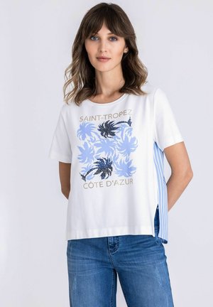 Vrouw draagt een wit shirt met korte mouwen met blauwe palmbomenprints en de tekst "Saint-Tropez Côte d'Azur", gecombineerd met een blauwe spijkerbroek, neutrale achtergrond.
