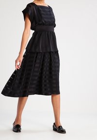 Černý dvoudílný outfit zahrnující peplum top s krátkými rukávy a plisovanou sukni, oba z průhledné látky s vertikálními pruhy.