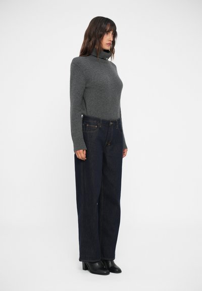 Pullover grigio con colletto a dolcevita dalla texture morbida abbinato a jeans a gamba larga blu scuro. La modella è in piedi di lato, indossando stivaletti neri con tacco.