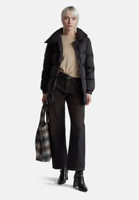 Černá péřová bunda s vysokým límcem, béžový svetr, tmavé široké džíny a černé kožené boty na podpatku. V ruce má kostkovanou tote tašku.