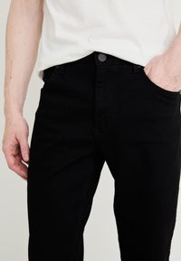 Bărbat purtând blugi negri și o cămașă albă, o mână în buzunar și cealaltă relaxată pe lângă corp.