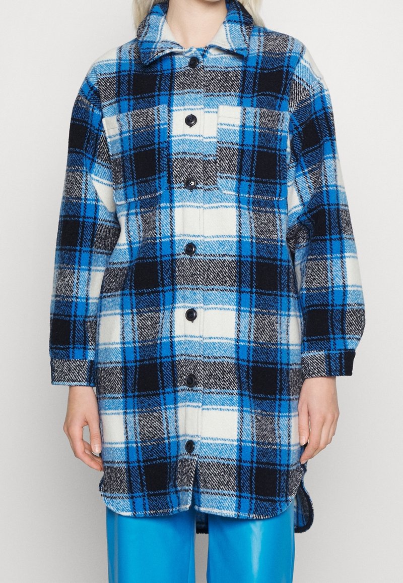 Giacca camicia a quadri in blu, nero e bianco. Realizzata in tessuto strutturato con chiusura a bottoni, colletto appuntito e una tasca sul petto.