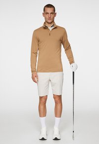 Beżowa koszulka golfowa z długim rękawem i zamkiem przy kołnierzu, zestawiona z jasnymi szortami. Model trzyma kij golfowy, nosząc białe buty i skarpetki.