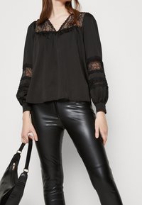 Svart blus med spetsdetaljer, med V-ringning och långa ärmar. Parat med svarta läderbyxor och håller en svart handväska.