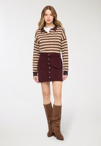 Giovane donna che indossa un polo a maniche lunghe a righe marroni, gonna bordeaux con bottoni sul davanti e stivali marroni in suede alti fino al ginocchio, in piedi su sfondo bianco.