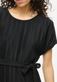 Svart plisserad klänning med vid halsringning, korta ärmar och ett bälte i tyg. Mjuk, slät textur med vertikala ränder genom hela plagget.