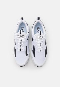 Bílé sportovní boty s černými abstraktními vzory, texturovaný síťovaný svršek, bílé tkaničky a logo na jazyku a patě.