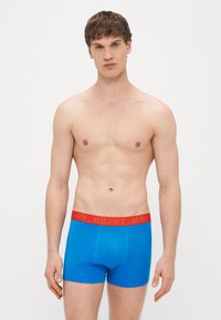Cuecas tipo trunk de algodão azul com uma cintura elástica vermelha com a palavra "HAPPY" impressa repetidamente. O design é simples e ajustado.