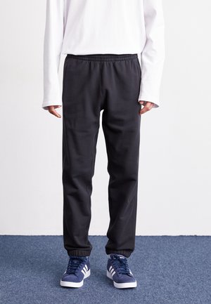 Osoba ubrana w czarne spodnie dresowe z elastycznym pasem, białą koszulę z długim rękawem i niebieskie buty sportowe Adidas stoi na niebieskim dywanie.