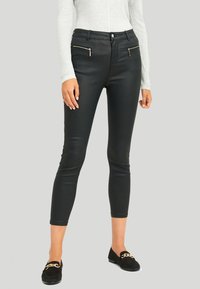 Svarta fusk läder tights med framfickor med dragkedja, slim fit och kort längd, kombinerade med svarta loafers och en grå tröja.