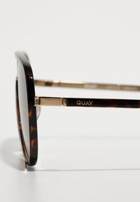 Gafas de sol cuadradas con una montura de carey y acentos metálicos en dorado. Presenta un logo impreso en dorado en la patilla. Lentes oscuros.