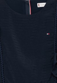 Námornícko modrá blúzka s horizontálnou rýhovanou textúrou, okrúhlym výstrihom a volánikovými detailmi. Obsahuje malé logo Tommy Hilfiger v červenej, bielej a modrej farbe.