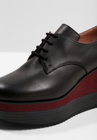 Chaussure à plateforme en cuir noir, à lacets, avec une épaisse semelle bordeaux et noire, des coutures visibles et une doublure intérieure beige.