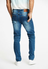 Pantalones vaqueros de mezclilla azul con un ajuste Slim, que presentan un parche de cuero marrón en la cintura, bolsillos traseros y áreas ligeramente deslavadas para dar textura.