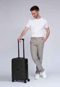 Man in een wit T-shirt en grijze broek staat met één hand in zijn zak en houdt een zwarte koffer met wielen vast aan de uitgetrokken handgreep.