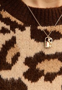 Colgante en forma de corazón con cerradura dorada sobre una cadena enlazada, descansando sobre un suéter de punto con patrón marrón y beige y acabado texturizado.