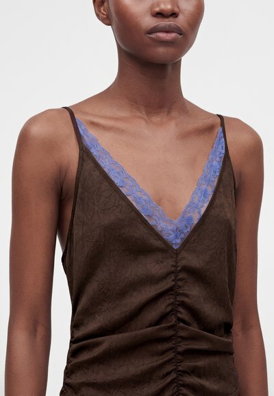 Femme portant un top marron sans manches froncé avec des fines bretelles et un décolleté en V dévoilant une bordure en dentelle bleue en dessous.