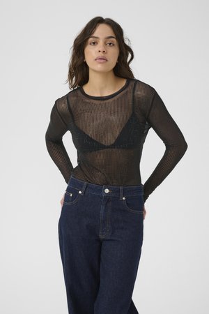 Vrouw met golvend haar, gekleed in een zwarte doorzichtige top met lange mouwen over een zwarte beha en donkerblauwe high-waisted jeans, staande tegen een witte achtergrond.