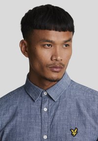 Lys blå button-up skjorte lavet af denimstof, med standard krave og broderet gult logo på brystet.