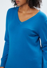 V-Ausschnitt Strickpullover in leuchtendem Blau, mit langen Ärmeln, geripptem Ausschnitt und einer weichen, glatten Textur ohne sichtbare Muster.