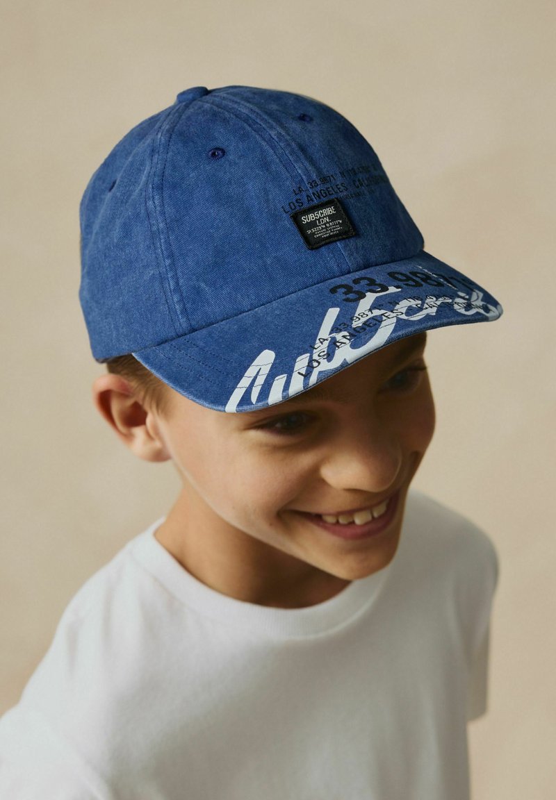 Casquette en coton bleue avec une visière courbée, comportant un texte graphique blanc et une étiquette noire à l'avant. Tissu texturé et détails de couture visibles.