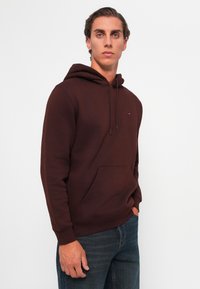 Brown hooded sweatshirt with a front pocket, made of soft fabric. Features drawstrings and a small logo on the chest. Worn with blue jeans.