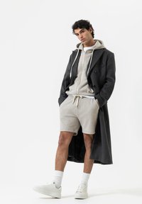 Manteau long gris sur un sweat à capuche gris clair avec cordons, associé à un short gris et des baskets blanches. Textures lisses, design minimal.