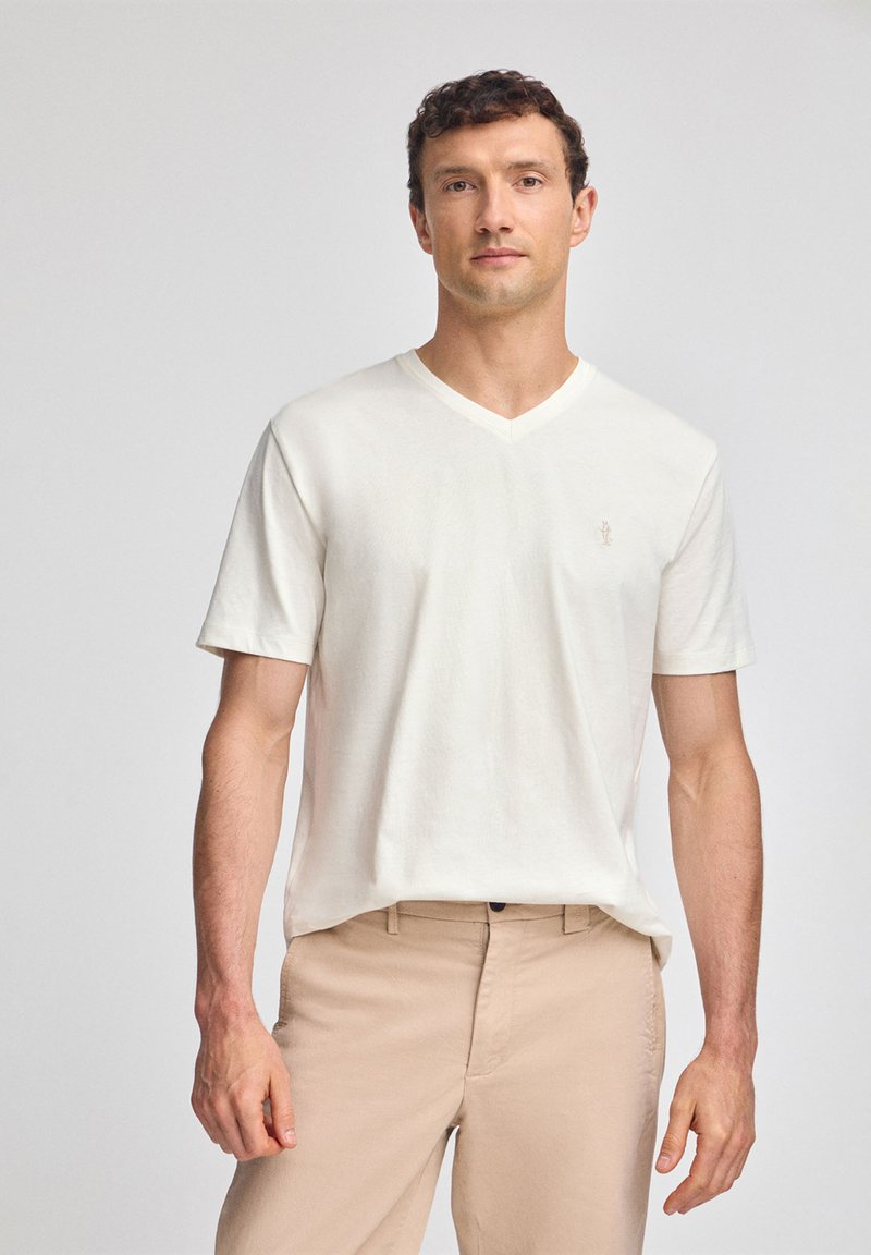 T-shirt bianca con scollo a V realizzata in tessuto morbido, con maniche corte e un piccolo logo ricamato sul petto sinistro. Indossata con pantaloni beige.