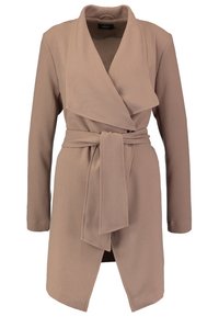 Beige naiset pitkähihaine kääntötakki, jossa on leveä kauluskääntö ja samaa kangasta oleva vyö, sidottuna vyötärölle.