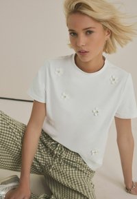 T-shirt blanche à col rond, à manches courtes, ornée de fleurs en tissu et de perles. Associée à un pantalon à carreaux verts.