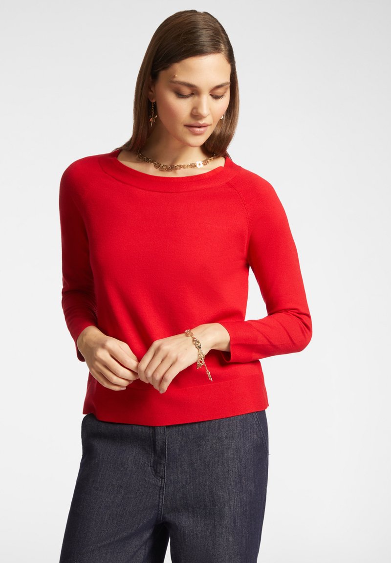 Maglietta rossa a maniche lunghe con scollatura rotonda e tessuto testurizzato. Indossata con pantaloni di denim scuri, arricchiti da dettagli in gioielleria dorata.