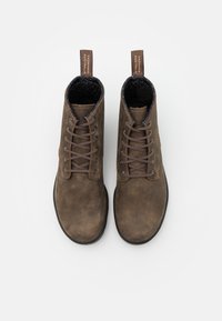 Botas de ante marrón hasta el tobillo con puntera redonda, ojales de metal y cordones marrones. También son visibles las lengüetas superiores y el forro negro texturizado.