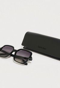 Černé čtvercové sluneční brýle s čočkami s přechodem, které mají silný rámeček a jemné značení. Součástí je také černé ochranné pouzdro.