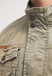 Jasno khaki kurtka z kołnierzem stójkowym, dwoma metalowymi guzikami, kieszenią na piersi oraz teksturowanym materiałem z widocznymi przeszyciami.
