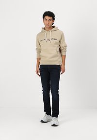 Μπεζ φούτερ με κορδόνια και μπροστινή τσέπη, που φέρει λογότυπο. Συνδυάζεται με σκούρα τζιν και λευκά αθλητικά παπούτσια. Απλός σχεδιασμός.