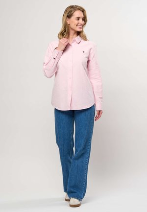 Smilende kvinde står iført en lyserød skjorte med knapper, blå vidde bukser og hvide sneakers mod en ensfarvet baggrund.