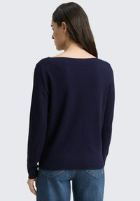 Frau, die mit dem Rücken zur Kamera steht, trägt einen navyblauen Langarmpullover und blaue Jeans vor einem schlichten hellen Hintergrund.