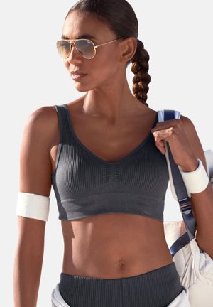 Woman with braided hair wearing sunglasses, dark ribbed sports bra and leggings, white wrist and arm bands, carrying a white gym bag.