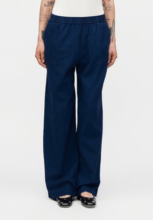 Άτομο που φοράει μπλε σκούρο παντελόνι με φαρδιά μπατζάκια και ελαστική μέση, καθώς και μαύρα παπούτσια με ασημένια καρφιά, με ορατά τατουάζ στα χέρια στο πλάι.