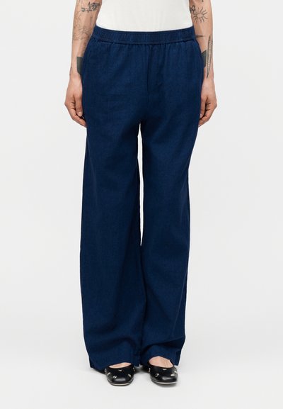 Personne portant un pantalon large bleu foncé avec une taille élastique et des ballerines noires avec des clous argentés, bras avec des tatouages visibles sur les côtés.
