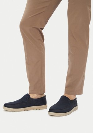 Granatowe buty wsuwane o zamszowej teksturze, z gumową podeszwą i beżowymi akcentami, w połączeniu z jasnobrązowymi, dopasowanymi spodniami.