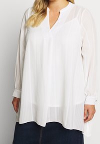 Blusa bianca a maniche lunghe con scollatura a V, caratterizzata da righe verticali, vestibilità ampia e polsini con bottone. Tessuto morbido e leggero.