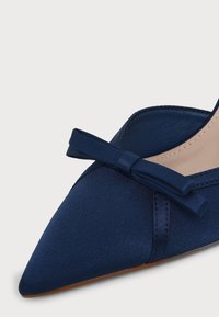 Chaussure plate en satin bleu marine à bout pointu avec un petit nœud assorti sur le dessus et une doublure intérieure beige.