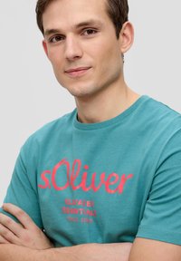 Turquoise t-shirt met een ronde halslijn met vetgedrukte rode tekst "s.Oliver" en kleinere tekst eronder. Soepele katoenen stof, korte mouwen.