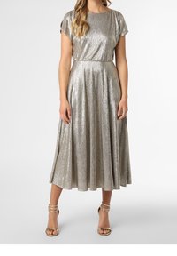 Femme portant une robe midi argentée métallique à manches courtes, avec une taille cintrée et des sandales à talons hauts dorées à lanières.