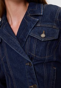 Tume teksajakk, millel on kohandatud disain, silmapaistev õmblus, suur krae ja rinnatasku ümmarguse metallnupuga.