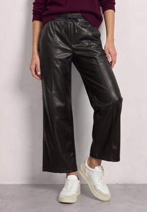Une personne porte un pantalon large noir en simili cuir avec une taille à cordon, associé à un pull bordeaux et des baskets blanches.