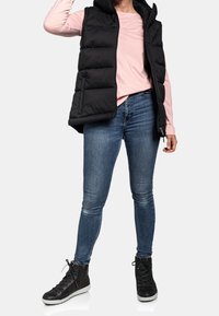 Czarna kamizelka puchowa z kapturem, noszona na jasnoróżowej bluzce z długim rękawem, w komplecie z niebieskimi rurkami i czarnymi trampkami za kostkę.